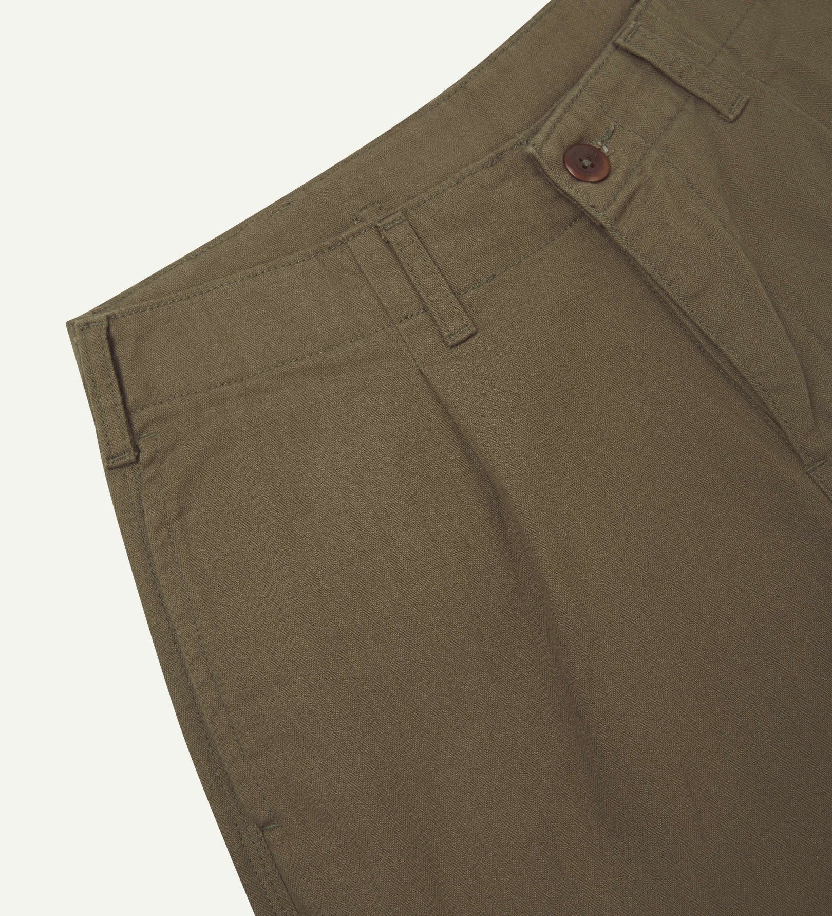 Close up, angled view of Uskees barrel leg pants waist area. Clear view of front pocket, belt loops, Corozo button fastening and organic cotton herringbone fabric.