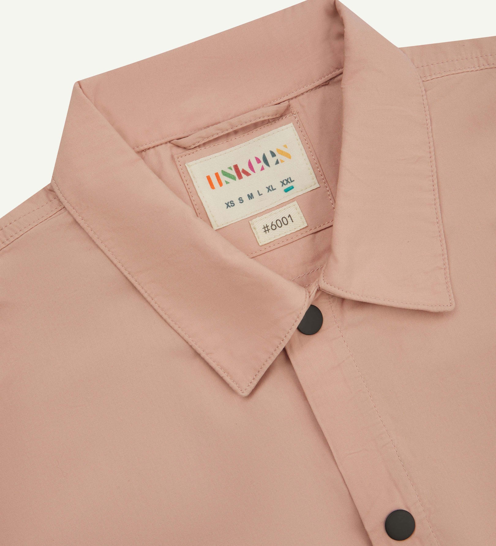 Front close-up view of the collar, popper buttons and Uskees branding label of the dusty pink 6001 lightweight overshirt.