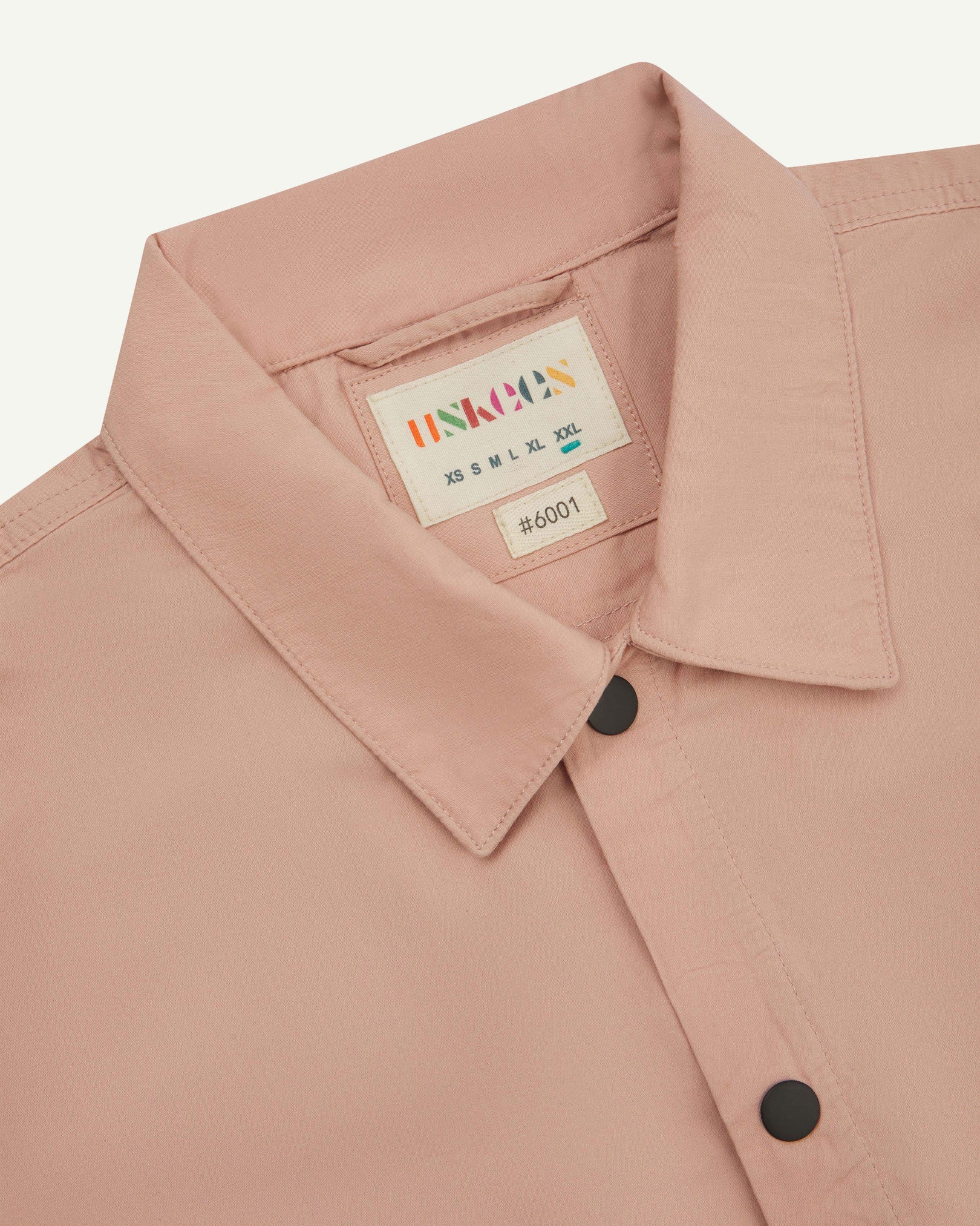 Front close-up view of the collar, popper buttons and Uskees branding label of the dusty pink 6001 lightweight overshirt.