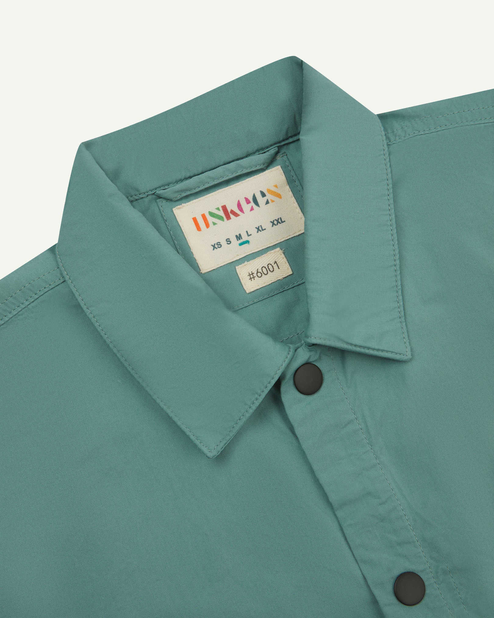 Front close-up view of the collar, popper buttons and Uskees branding label of the blue-green eucalyptus 6001 lightweight overshirt.