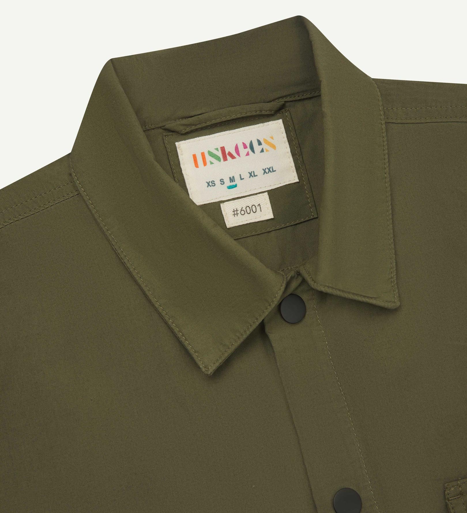 Front close-up view of the collar, popper buttons and Uskees branding label of the olive-green 6001 lightweight overshirt.