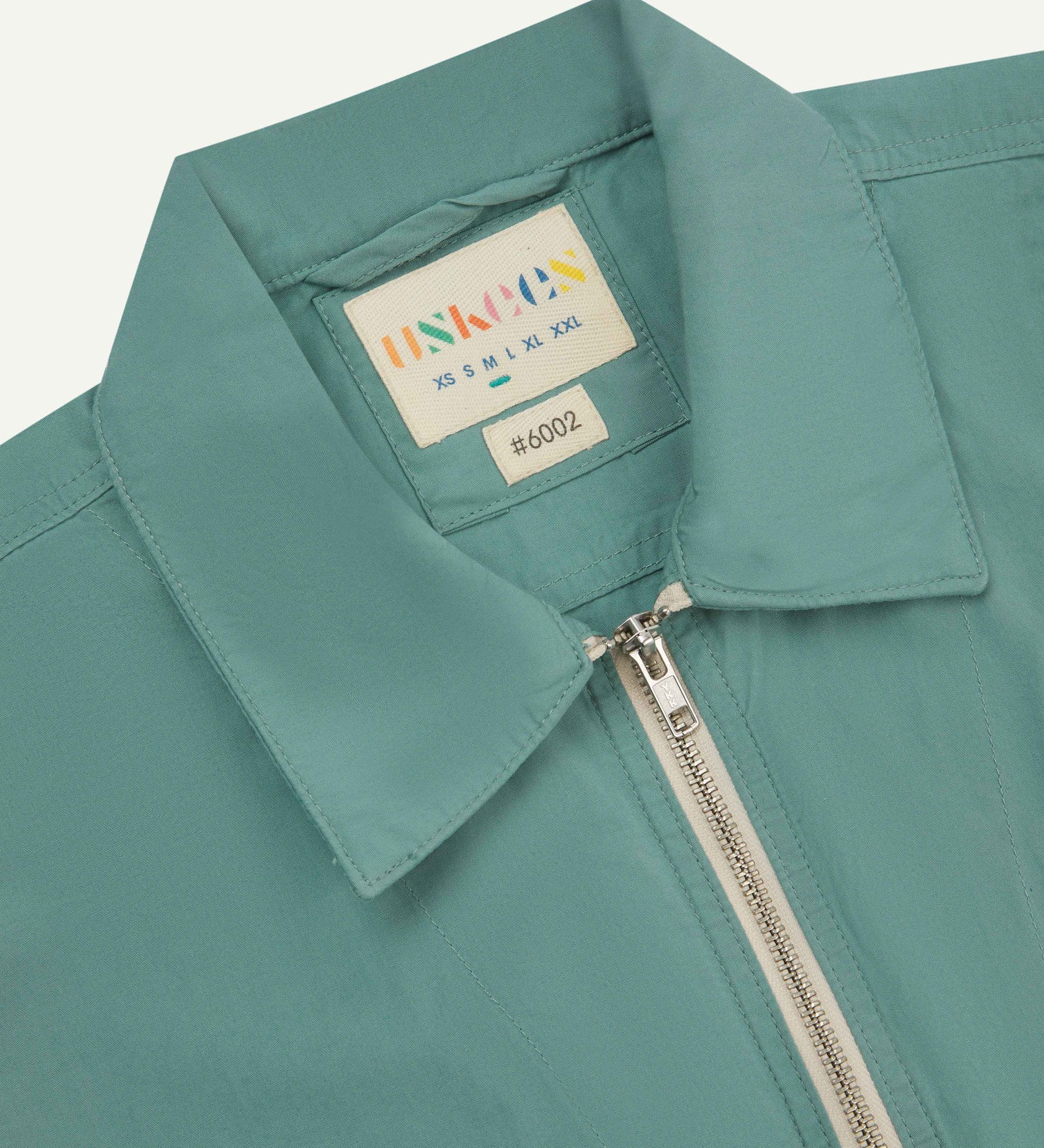 Front close-up view of the collar, zip detail and Uskees branding label of the the blue-green 'eucalyptus' coloured 6002 lightweight jacket.