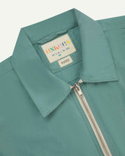 Front close-up view of the collar, zip detail and Uskees branding label of the the blue-green 'eucalyptus' coloured 6002 lightweight jacket.