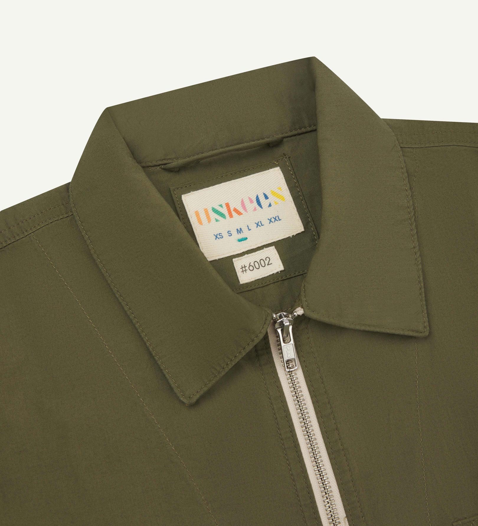 Front close-up view of the collar, zip detail and Uskees branding label of the olive-green 6002 lightweight jacket.