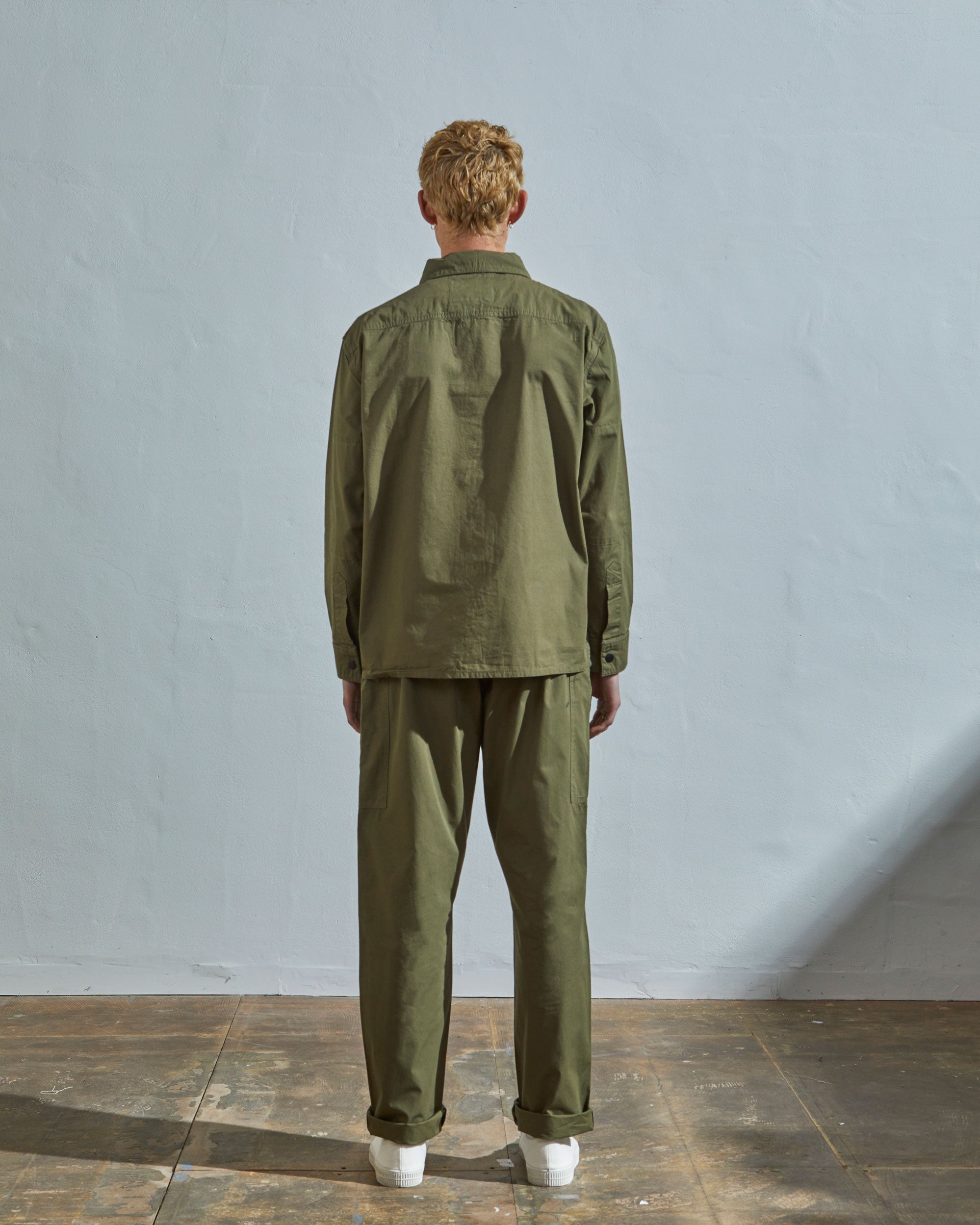 Full-length back view of model wearing Uskees #6002 olive lightweight zip front jacket, showing reinforced elbows and button sleeve placket.