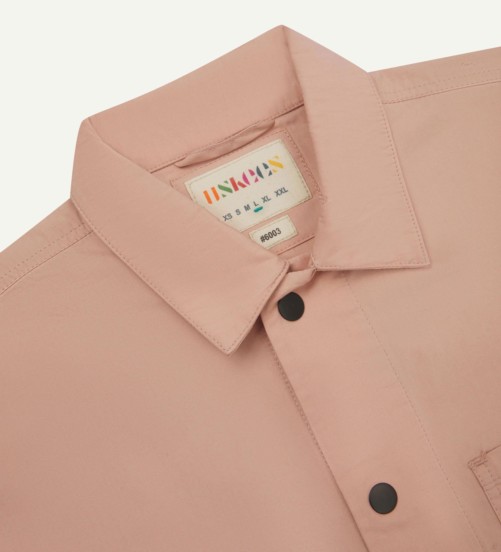 Close-up view of the #6003 reinforced shirt collar, showing weave of dusty pink organic cotton, contrast press studs and Uskees branding label.