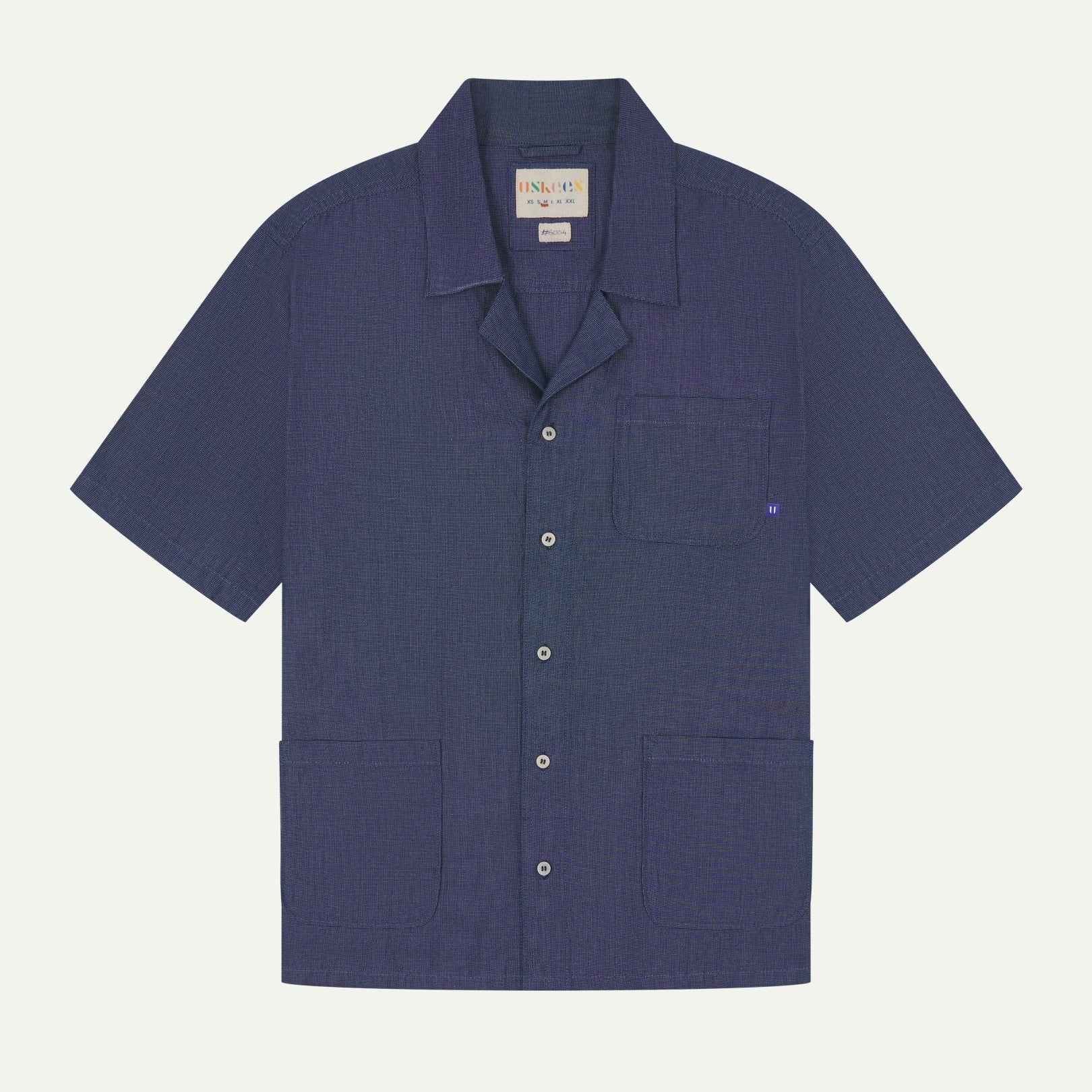 Flat front view of mid blue organic chambray Cuban utility shirt from Uskees. Clearly showing 3 front pockets and corozo button front.