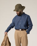 Bearded model wearing Uskees blue chambray oxford shirt, khaki trousers and green hat, holding a khaki blazer.