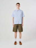 Full-length front view of model wearing Uskees Tasker buttoned short sleeve shirt with blue, gold and white striped cotton fabric. Paired with olive-green cargo shorts.
