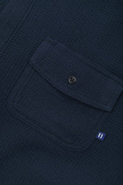 Angled mid-view of #6007 buttoned organic cotton waffle fabric short sleeve shirt in deep blue with focus on button down flap chest pocket and Uskees branding tag.