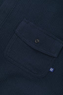 Angled mid-view of #6007 buttoned organic cotton waffle fabric short sleeve shirt in deep blue with focus on button down flap chest pocket and Uskees branding tag.