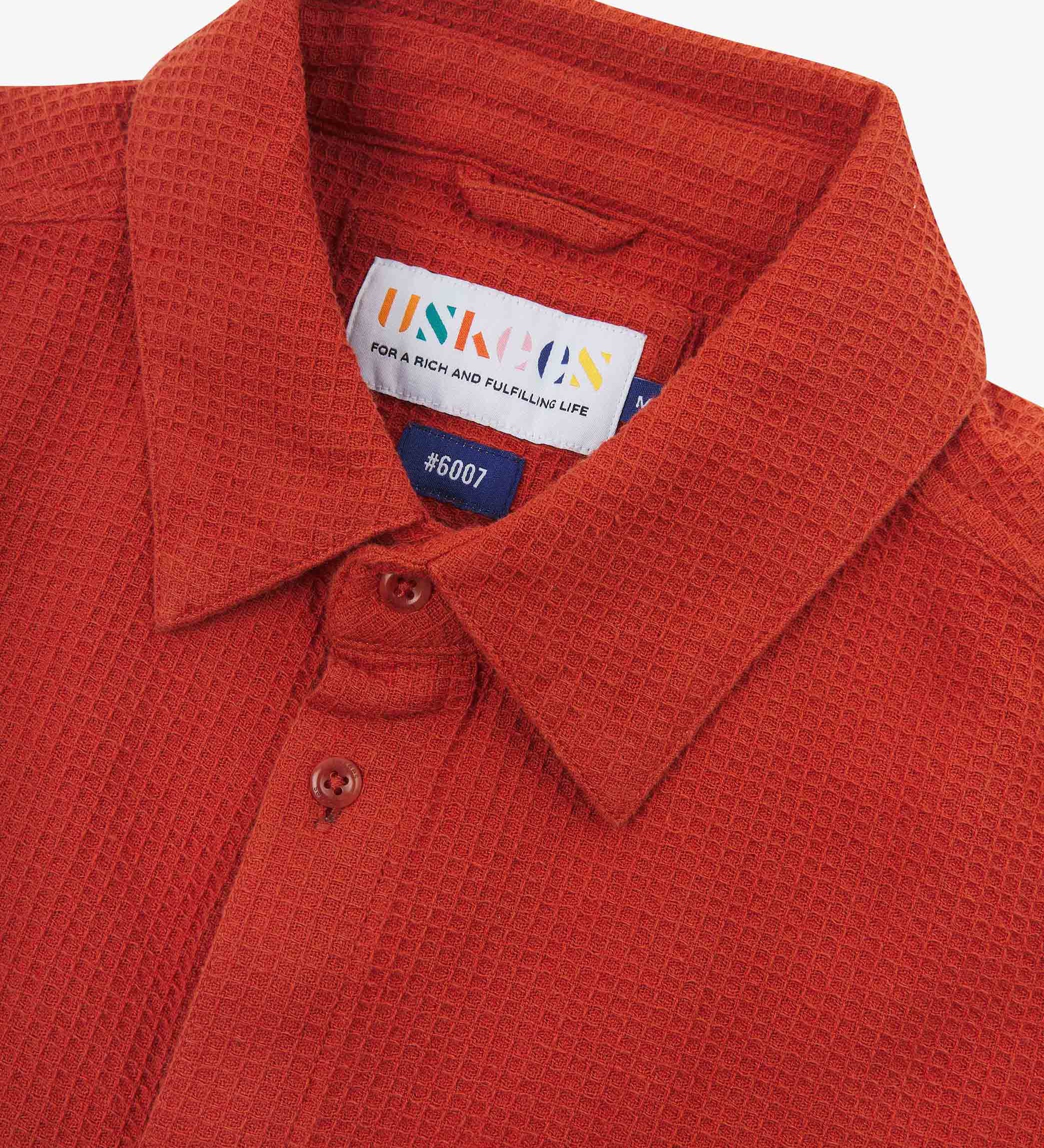 Close-up and angled front view of the 6007 Tasker shirt in rust-red, showing the waffle texture, collar, hanging loop and matching corozo buttons.