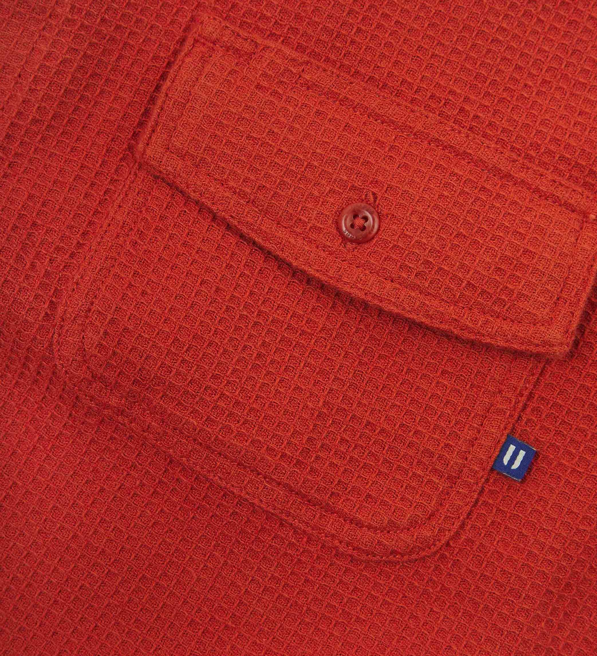 Angled mid-view of #6007 buttoned organic cotton waffle fabric short sleeve shirt in rust-red with focus on button down flap chest pocket and Uskees branding tag.