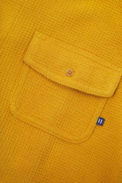 Angled mid-view of #6007 buttoned organic cotton waffle fabric short sleeve shirt in yellow with focus on button down flap chest pocket and Uskees branding tag.