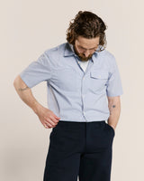 Man wearing an Uskees light blue short sleeve striped shirt and dark pants against a beige background.