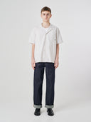 Full-length front view of model wearing an Uskees off-white Cuban shirt paired with indigo raw orta Uskees jeans.