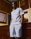 Male model wearing an Uskees light blue striped shirt and shorts in an indoor setting with wooden walls.