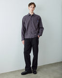 A male model wearing a blue plaid long-sleeve button-down shirt, black trousers, and black leather loafers, standing against a light gray wall.