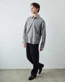 A man wearing an Uskees gray and white vertically striped long-sleeve button-down shirt with black trousers and black loafers, standing against a plain light gray wall.