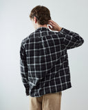 A man shown from behind wearing a charcoal and white plaid shirt and khaki pants, standing against a light gray studio background.