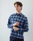 A male model wearing an Uskees blue and white plaid button-down shirt and dark navy pants, standing against a light gray studio background.