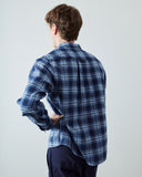 A man viewed from behind wearing a blue and white plaid button-down shirt and dark navy pants, standing against a light gray studio background.