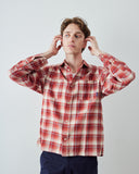 A man wearing a red and beige plaid long-sleeve button-up shirt and blue trousers, posing with his hands near his ears against a light gray studio background.