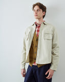A male model wearing an Uskees light beige overshirt over a brown knit cardigan and red plaid shirt, paired with navy trousers, standing against a plain light gray wall.