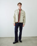 A man stands in a studio wearing a light beige overshirt over a red and white plaid shirt, navy blue trousers, and black leather shoes against a plain white wall and concrete floor.