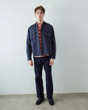A male model stands against a plain light gray wall wearing an Uskees navy blue overshirt over a red and blue plaid shirt, navy trousers, and black shoes.