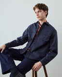 A male model wearing an Uskees navy blue utility jacket and matching trousers, layered over a red plaid shirt, sitting on a wooden stool against a light gray studio background.