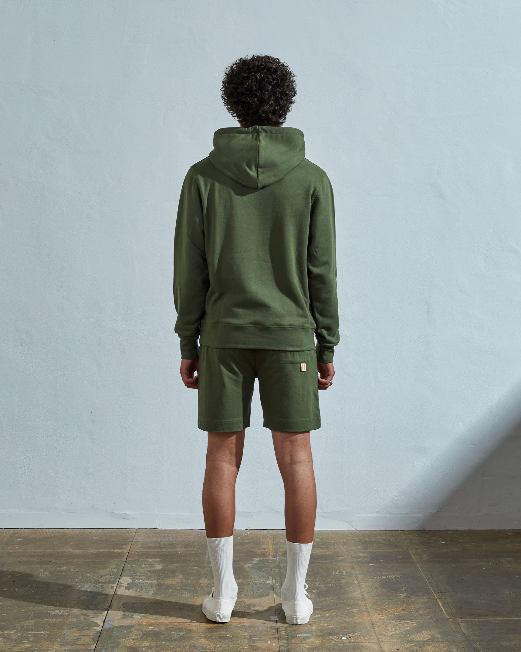 Full-length back view of model wearing coriander green, relaxed cut organic cotton #7004 jersey sweater. Showing hood, ribbed cuffs and hem.