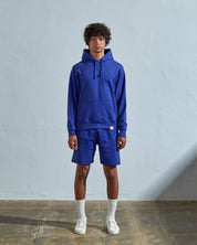 Full-length front view of model wearing 'ultra blue' organic cotton 7004 jersey hooded sweater with front pouch pocket. Paired with matching shorts