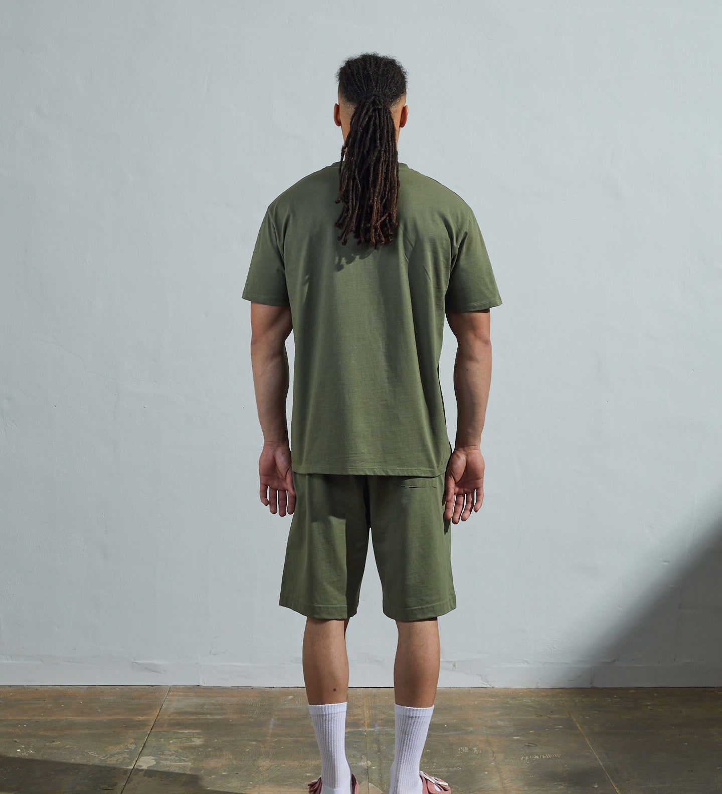 Full-length back view of model wearing army green, relaxed cut organic cotton #7006 jersey T-shirt by Uskees paired with matching #7007 shorts.