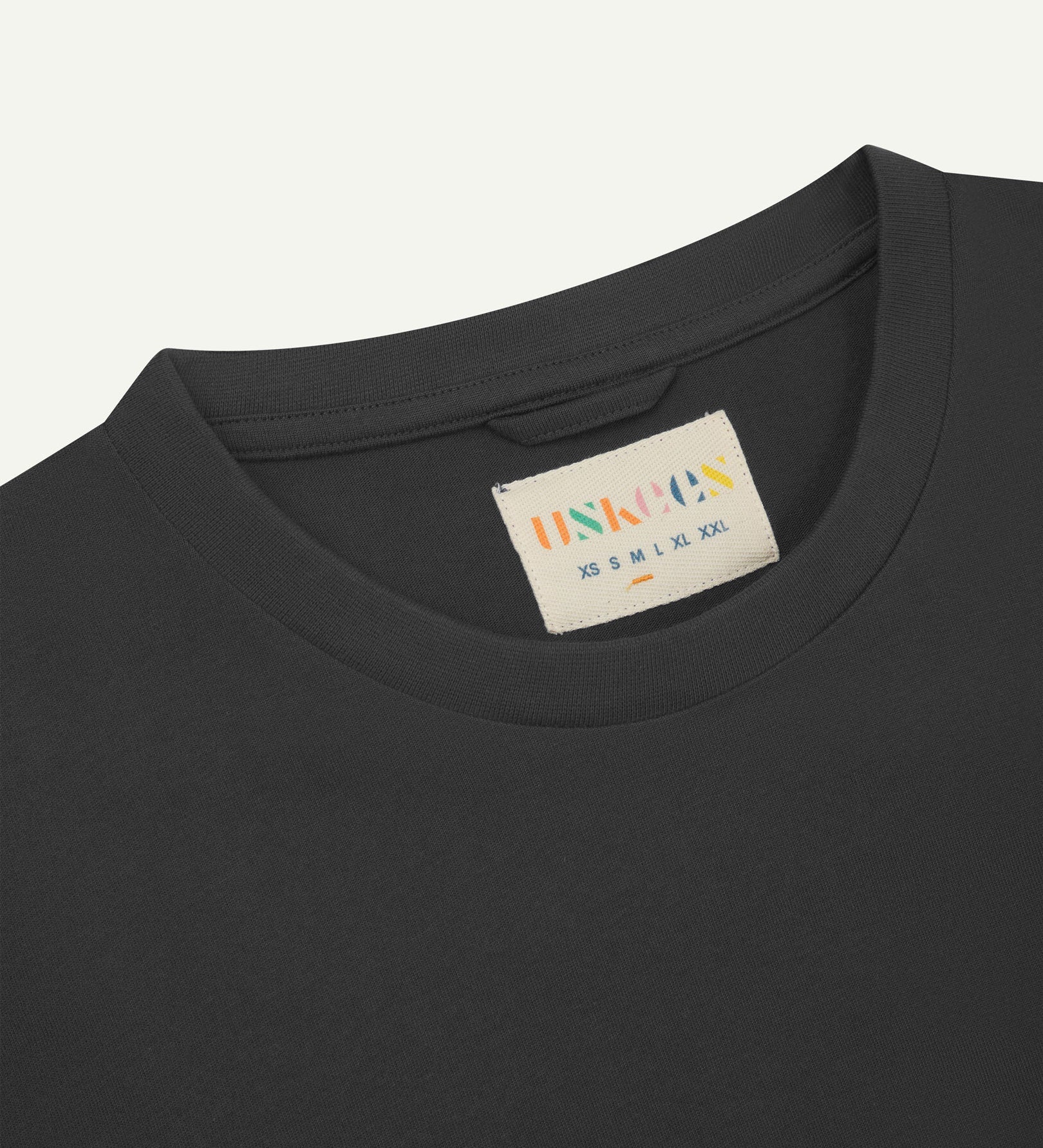 Neckline close-up of Uskees faded black organic cotton T-shirt showing hanging loop and branding label.