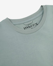 Neckline close-up of Uskees green-grey 'steel' coloured organic cotton T-shirt showing hanging loop and branding stamp.