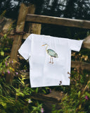 A white short-sleeve Uskees T-shirt  for men hanging on a rustic wooden fence outdoors, featuring a printed illustration of a heron on the chest.