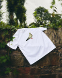 A white short-sleeve Uskees T-shirt with a printed pigeon graphic, draped over a rustic stone wall with green foliage and trees in the background.