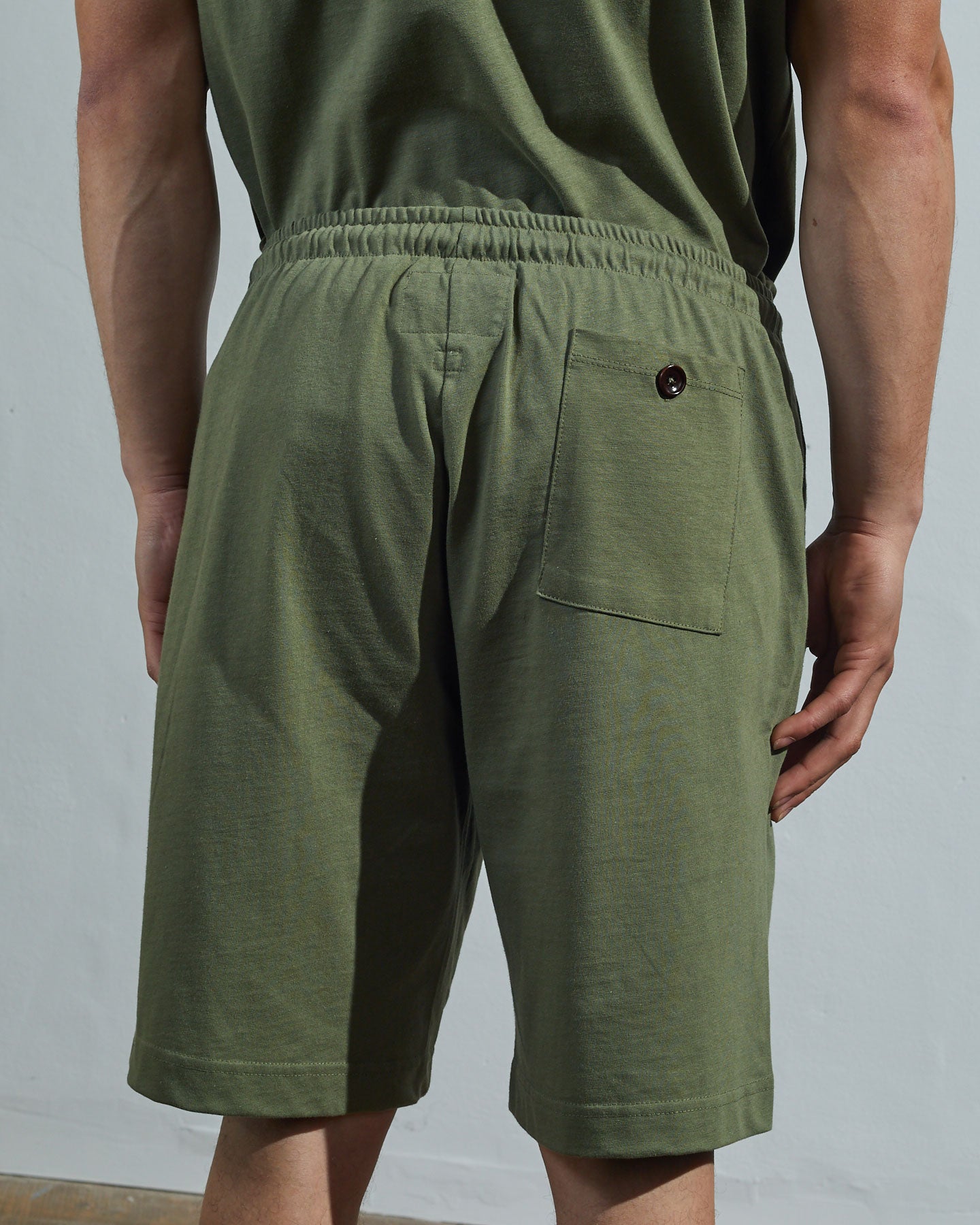 Back view of model wearing army green, relaxed cut organic cotton #7007 jersey shorts by Uskees with focus on back pocket with corozo button.