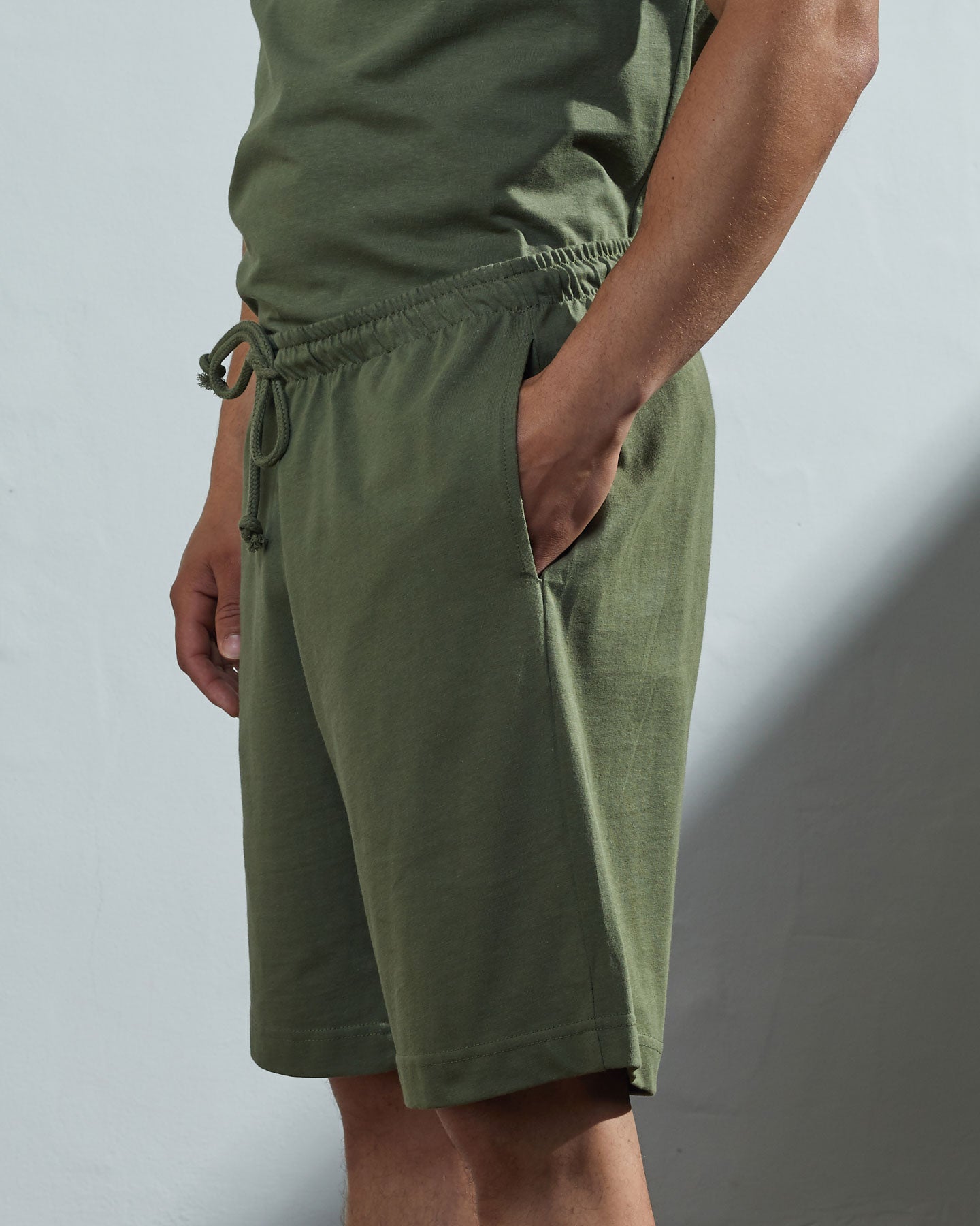 Side view of model wearing army green organic cotton #7007 jersey shorts by Uskees with hands in side pockets.