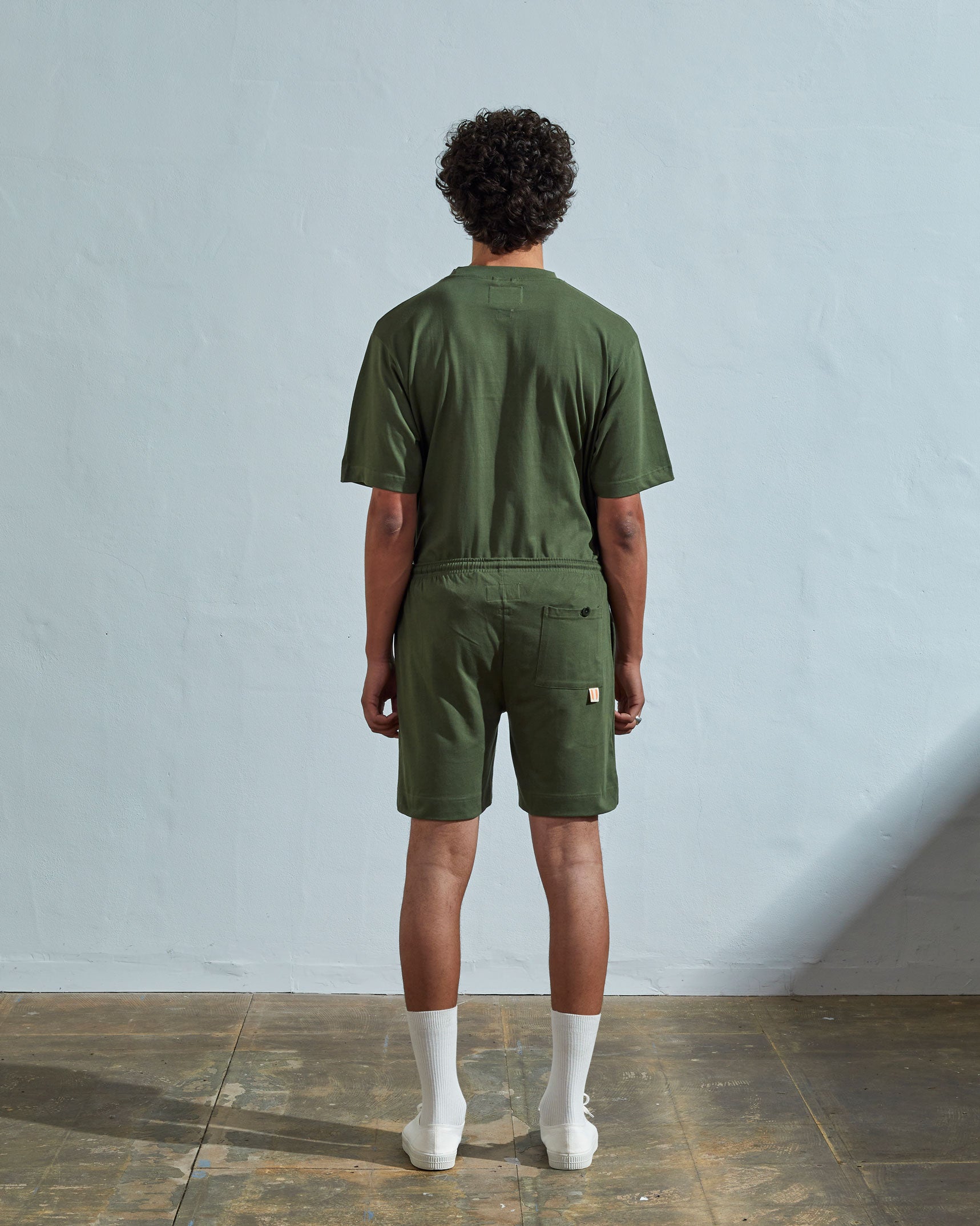 Back view of model wearing coriander-green, loose leg organic cotton jersey shorts by Uskees. Large front pockets visible.