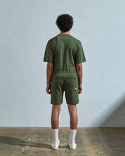 Back view of model wearing coriander-green, loose leg organic cotton jersey shorts by Uskees. Large front pockets visible.
