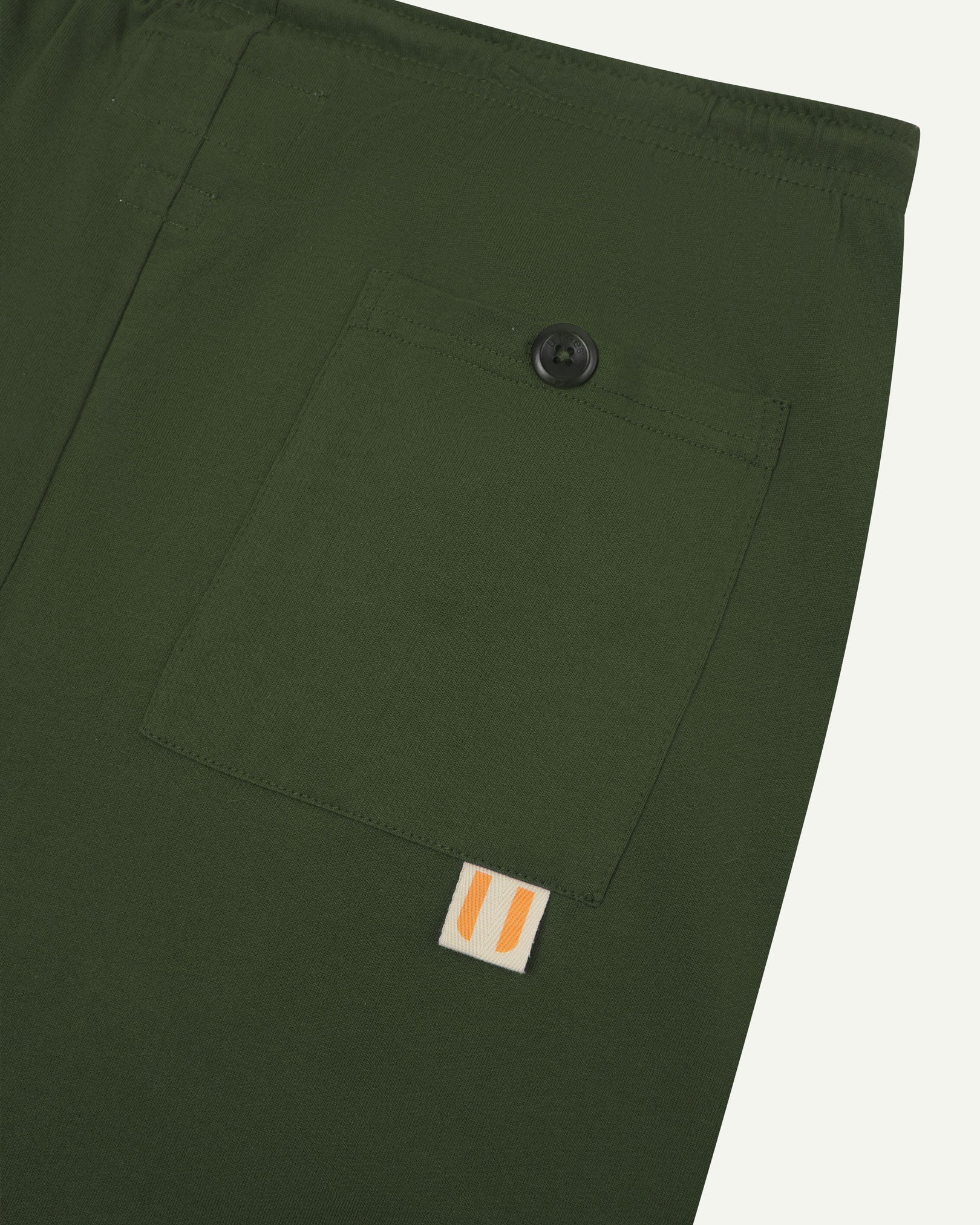 Close-up view of coriander-green organic cotton 7007 men's shorts against white background. Clear view of buttoned back pocket and Uskees logo label.