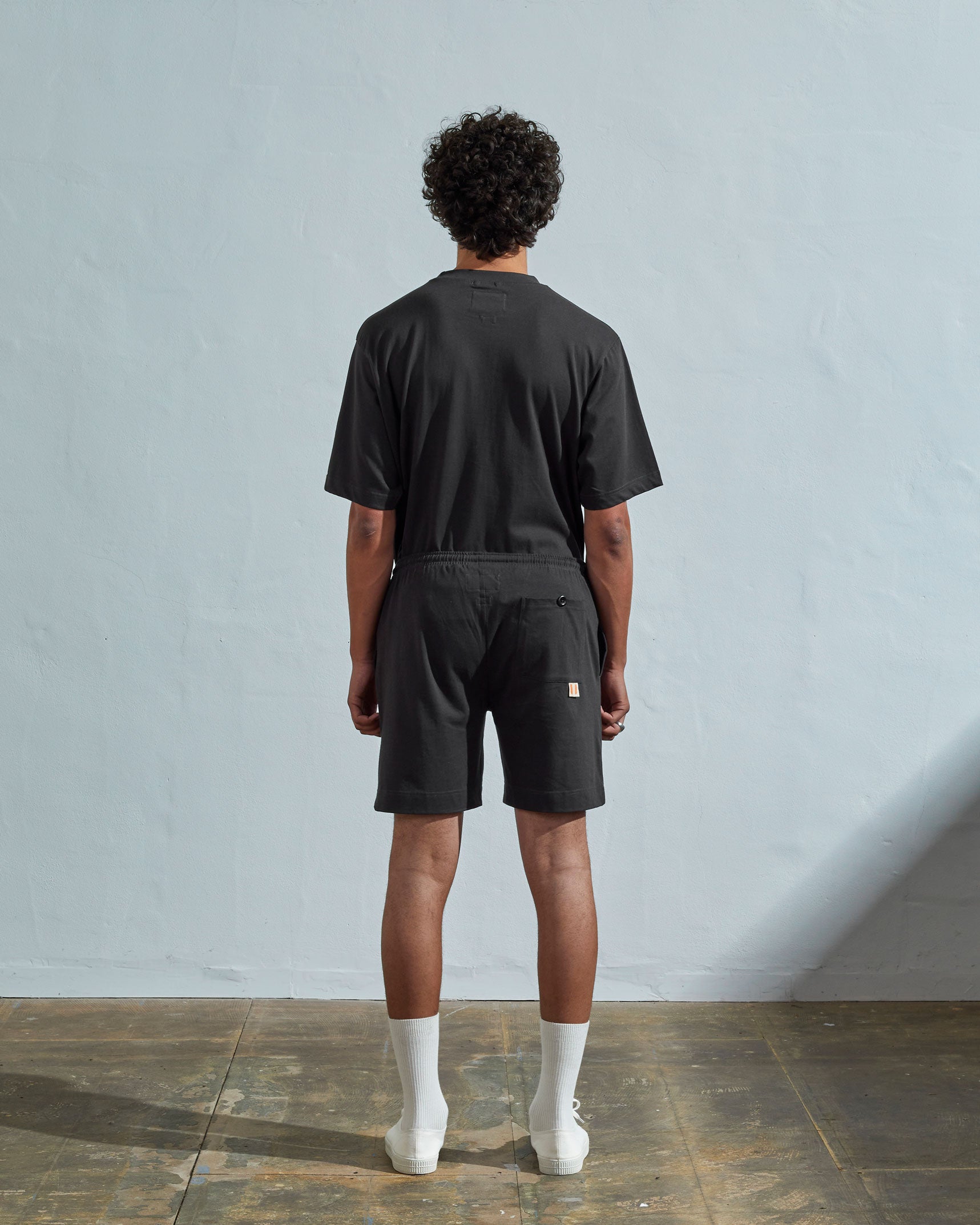 Back view of model wearing faded black, relaxed cut organic cotton #7007 jersey shorts by Uskees with focus on back pocket with corozo button.