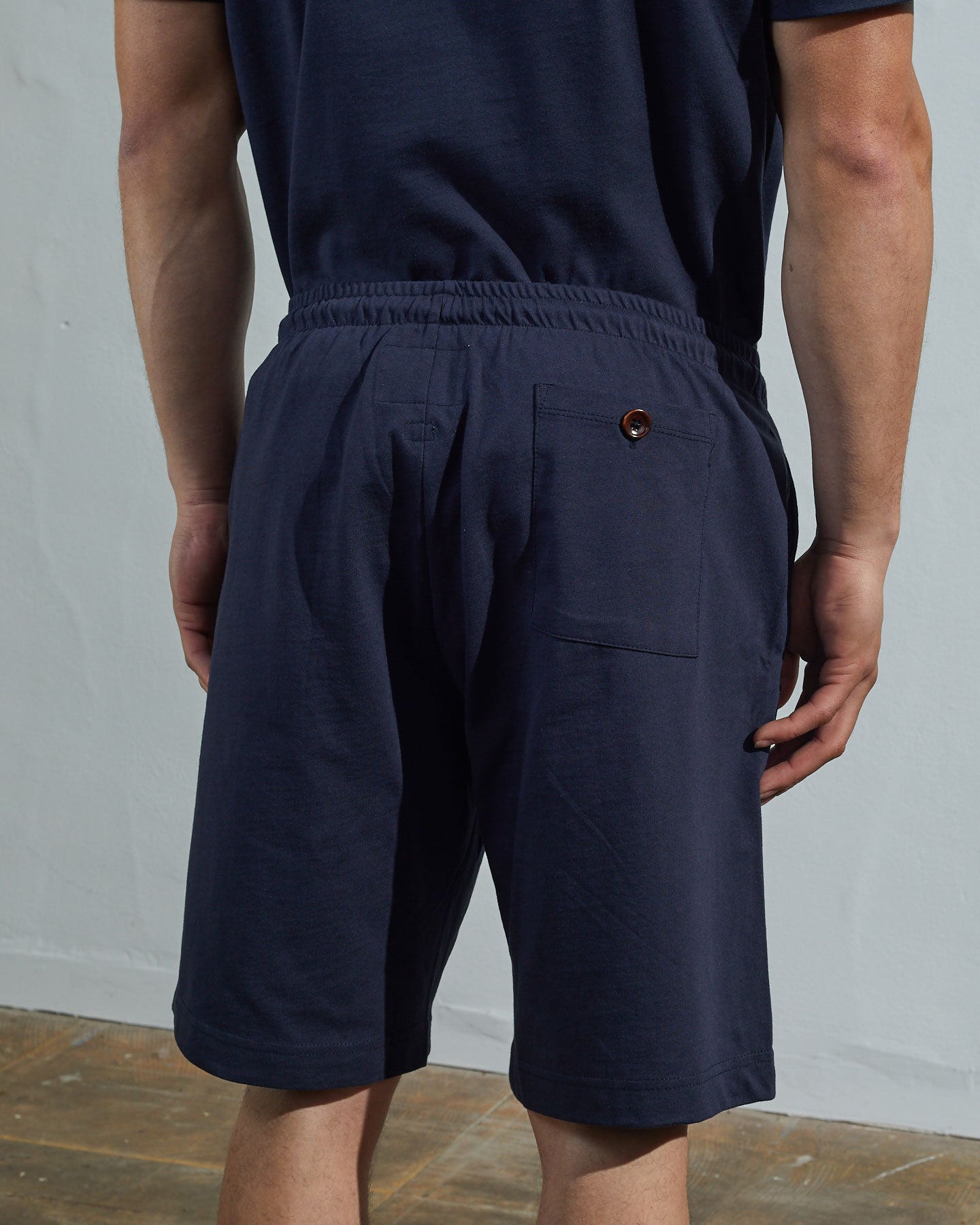 Back view of model wearing midnight blue, relaxed cut organic cotton #7007 jersey shorts by Uskees with focus on back pocket with corozo button.