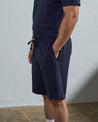 Side view of model wearing midnight blue organic cotton #7007 jersey shorts by Uskees with hands in side pockets.