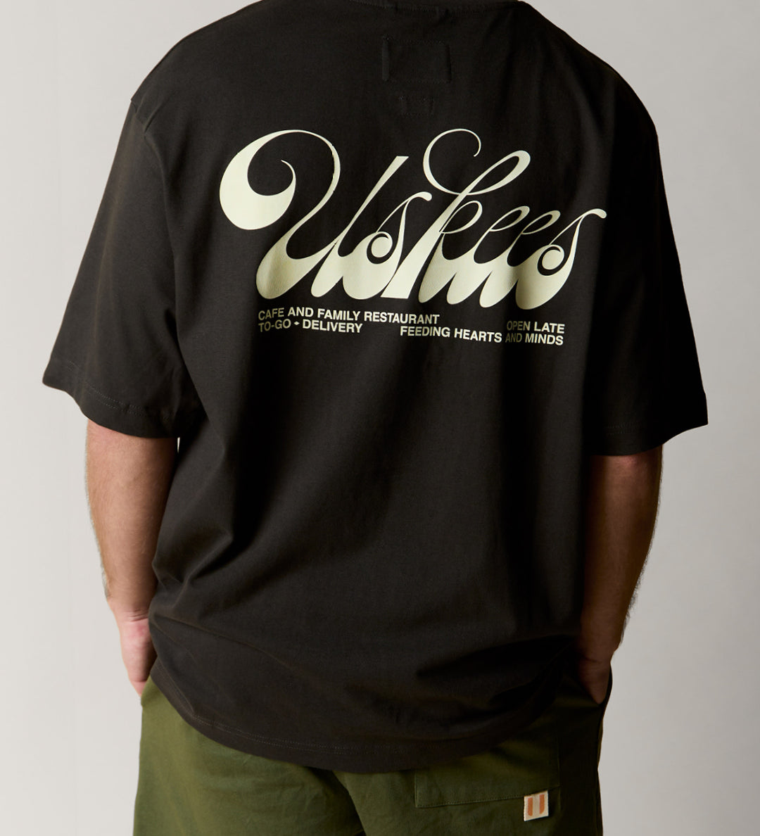 Reverse view of man wearing the uskees black men's 'diner' tee, demonstrating the oversized fit and the large, white 'diner' logo displayed across the back.