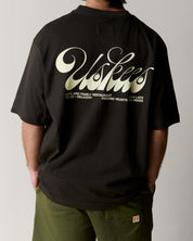 Reverse view of man wearing the uskees black men's 'diner' tee, demonstrating the oversized fit and the large, white 'diner' logo displayed across the back.