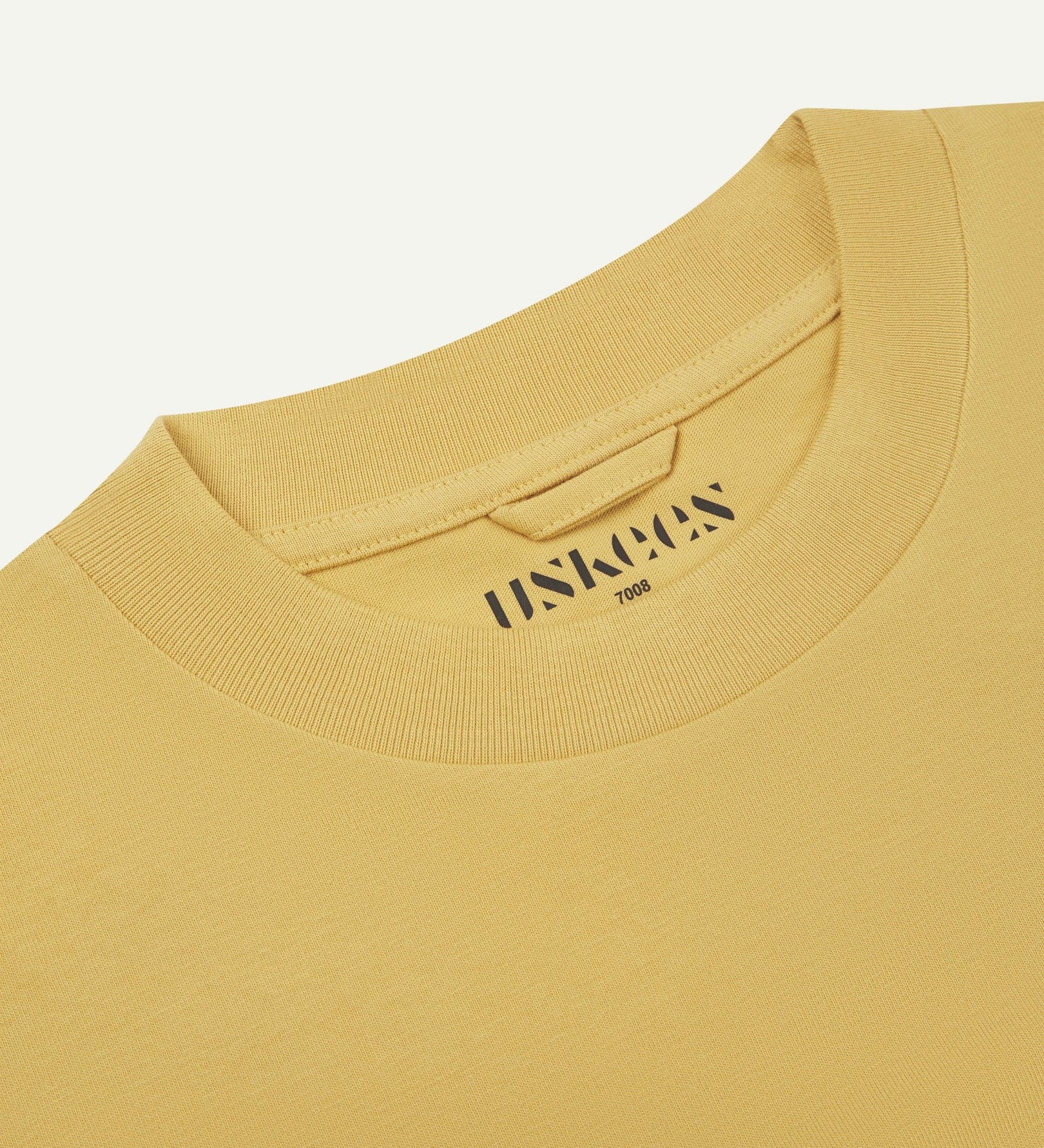 Neckline close-up of the oversized Uskees sandstone-yellow organic cotton T-shirt showing hanging loop and branding stamp.