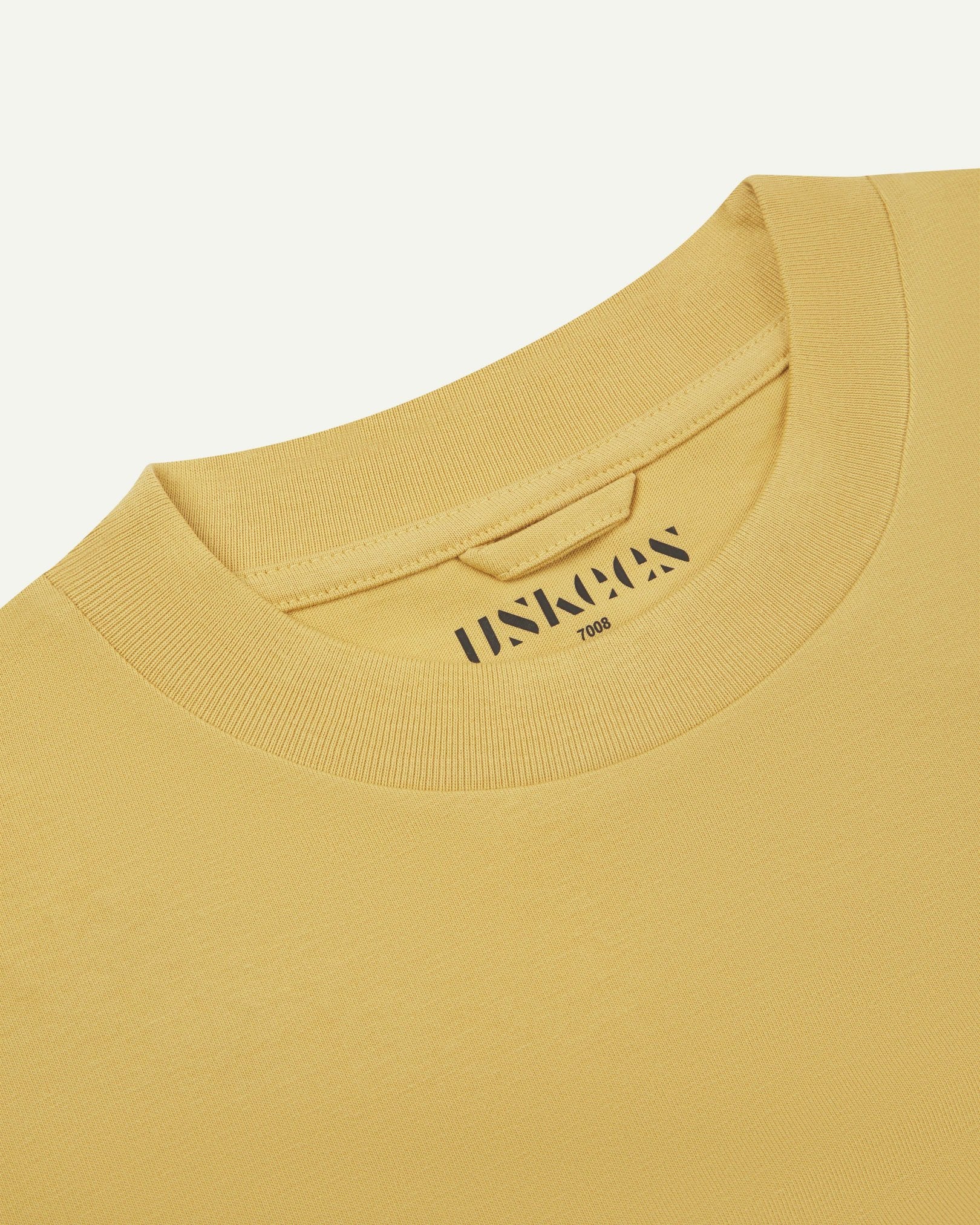 Neckline close-up of the oversized Uskees sandstone-yellow organic cotton T-shirt showing hanging loop and branding stamp.